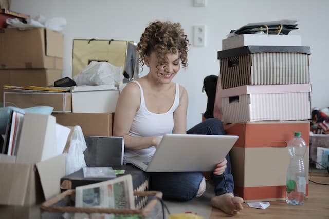 Woman posting stuff online for selling after decluttering her home while sitting on the floor surrounded by boxes
