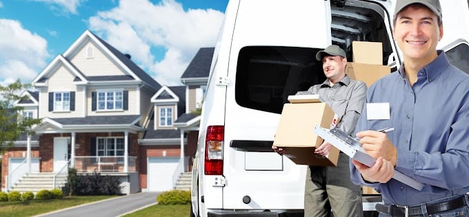 Two delivery workers with a van and packages stand in front of a house. One holds boxes while the other consults a clipboard, ensuring every detail is perfect. Choose the best for your next move and experience seamless service from start to finish.